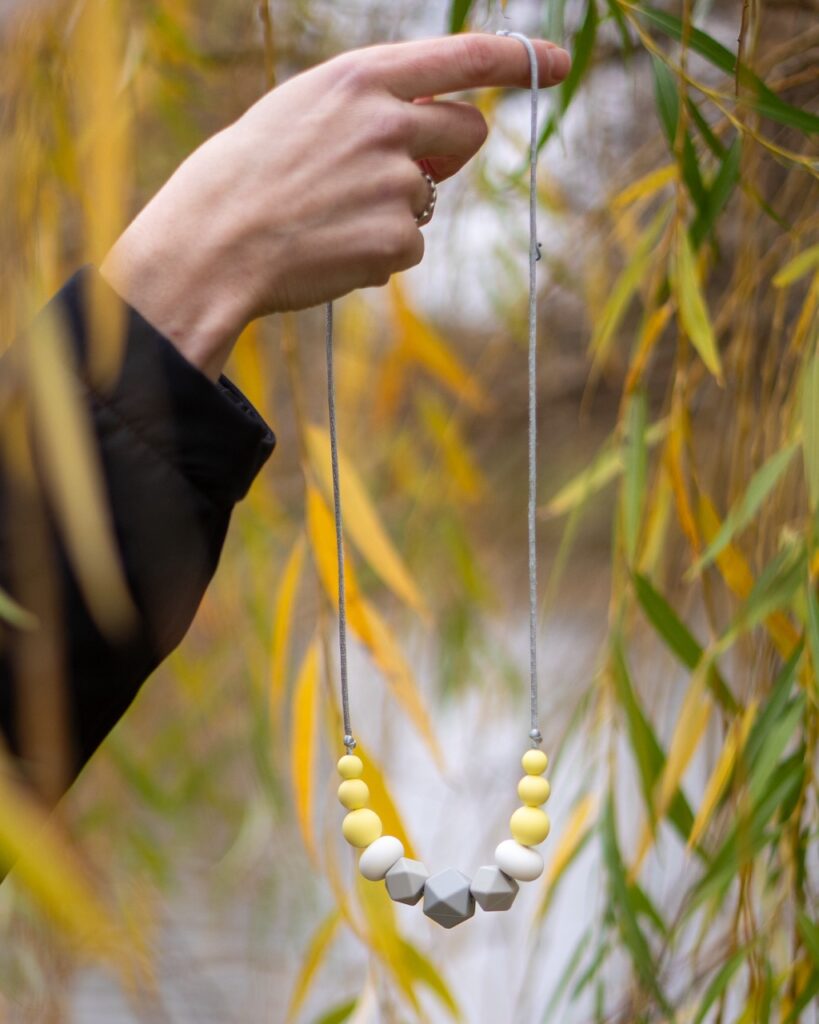 Collar de apego Lulumoon modelo Alba con piezas geométricas grises, blancas y amarillas colgando entre hojas de sauce.