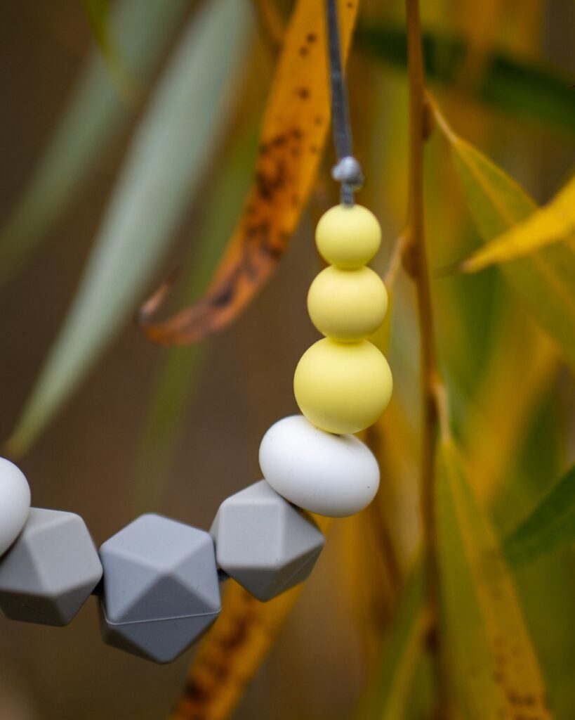 Collar de apego Lulumoon modelo Alba con piezas geométricas grises, blancas y amarillas colgando entre hojas de sauce.