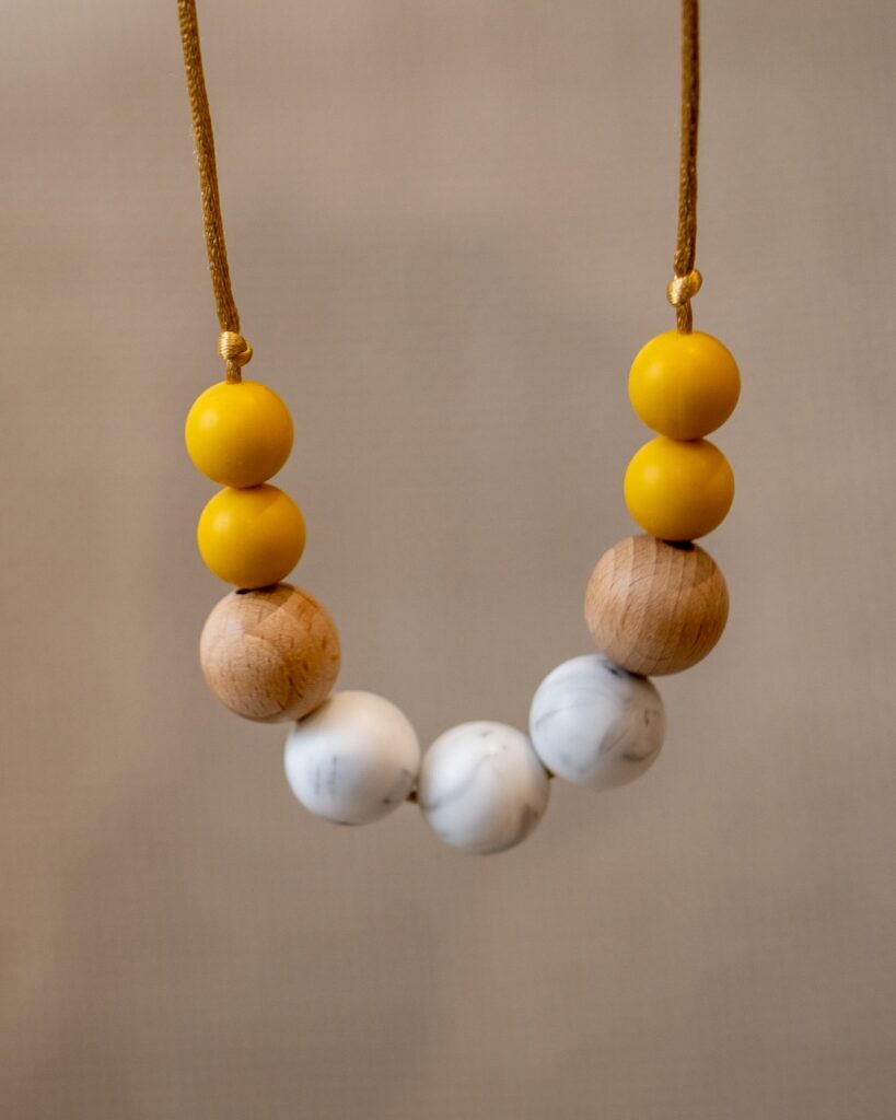 Collar de apego Sensory Lulumoon modelo Sol Nórdico estirado, mostrando las bolas en mostaza, mármol y madera sobre fondo neutro.