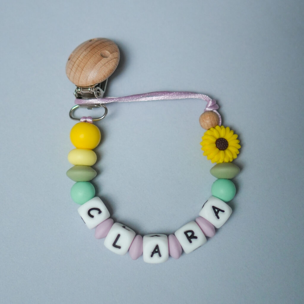 Chupetero con nombre CLARA y pieza central de girasol amarillo, con bolas de silicona en colores menta, lila y blanco.