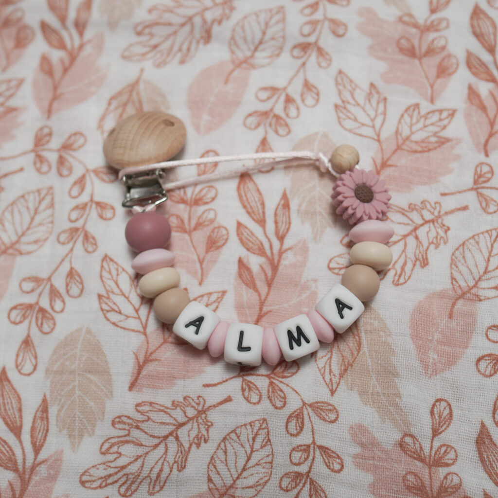 Texto Alternativo (Alt Text): Chupetero personalizado con el nombre ALMA y una margarita de silicona en tonos rosa terracota, polvo y beige sobre fondo de hojas.