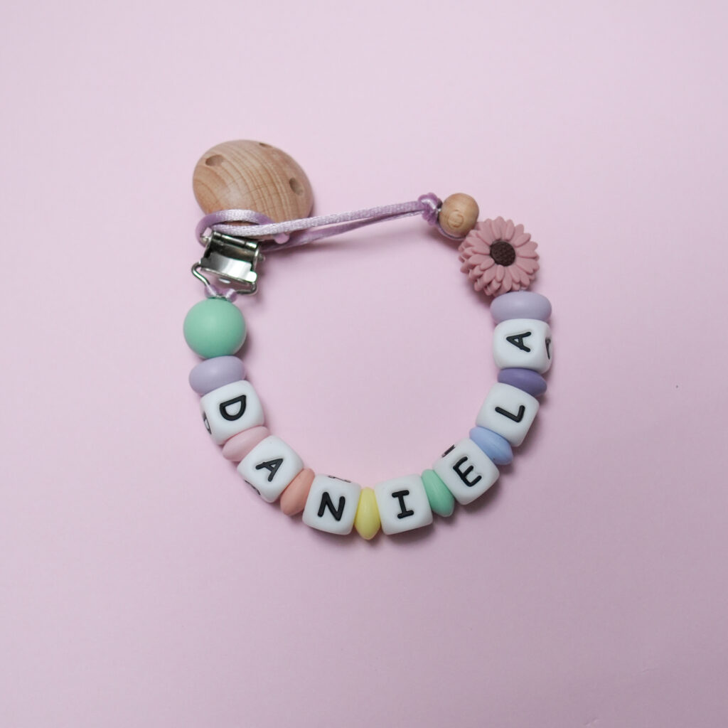 Sujeta-chupetes personalizado con el nombre DANIELA y margarita rosa empolvado, combinado con cuentas de silicona en colores pastel.