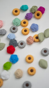 Chupetero de silicona con diseño de pétalos en degradado de colores arcoíris, sujeto por una mano y con clip de madera natural.