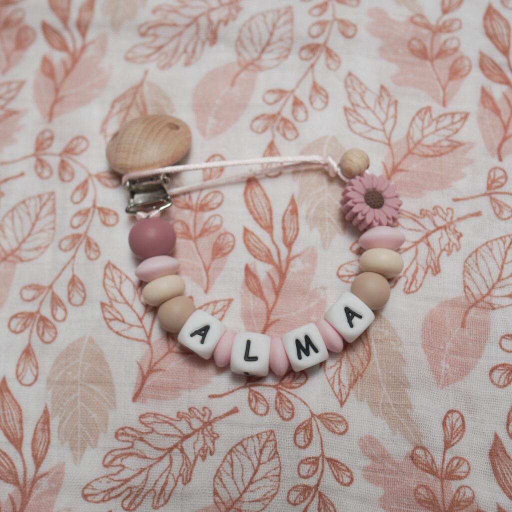 Chupetero personalizado con el nombre ALMA en tonos rosa empolvado y tierra, con pieza de silicona en forma de flor y clip de madera de haya natural.
