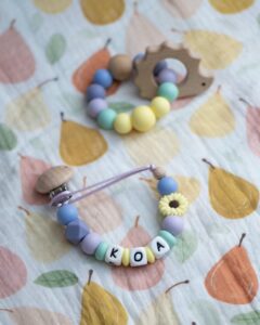 Chupetero personalizado con el nombre KOA, diseñado con cuentas de silicona en tonos lila, menta y amarillo pastel, incluyendo una pieza de girasol y clip de madera natural.