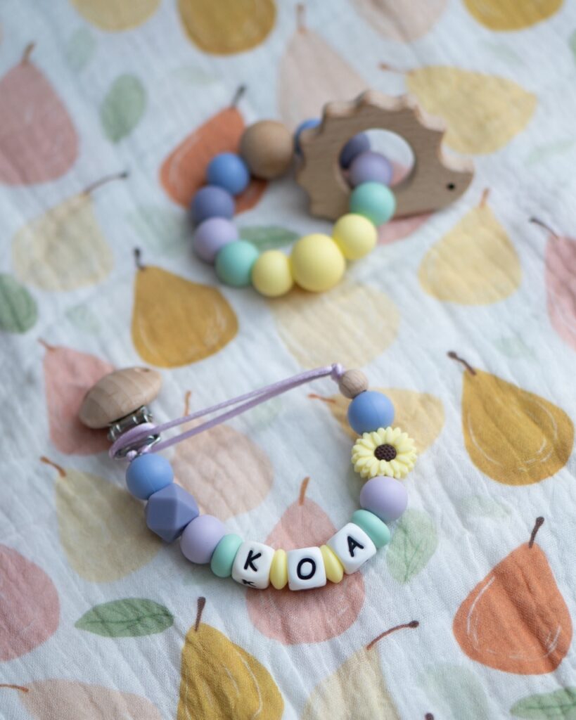 Chupetero personalizado con el nombre KOA, diseñado con cuentas de silicona en tonos lila, menta y amarillo pastel, incluyendo una pieza de girasol y clip de madera natural.