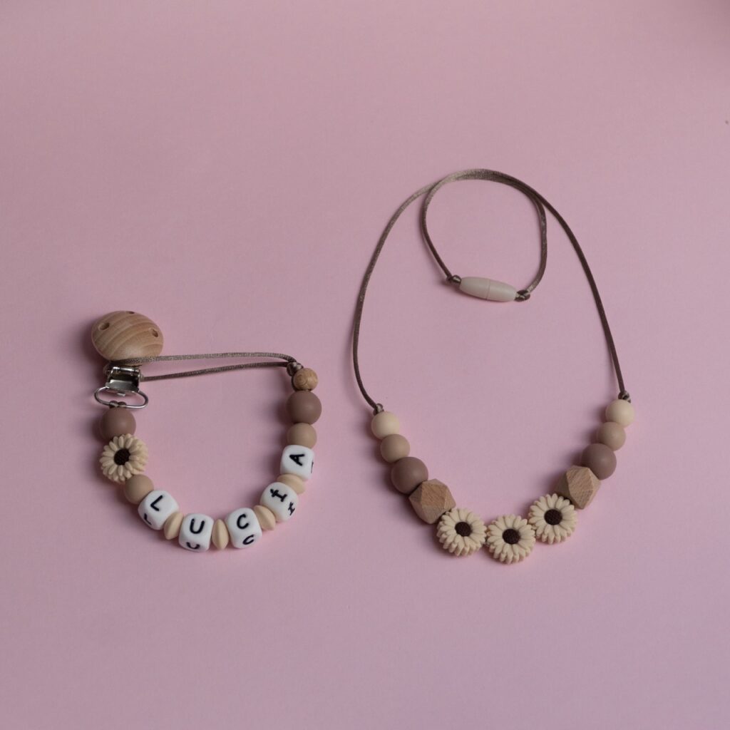 Conjunto de regalo para bebé compuesto por un chupetero personalizado con el nombre Lucía (incluyendo tilde) y un collar de lactancia a juego en tonos beige y tierra con detalles de girasoles.