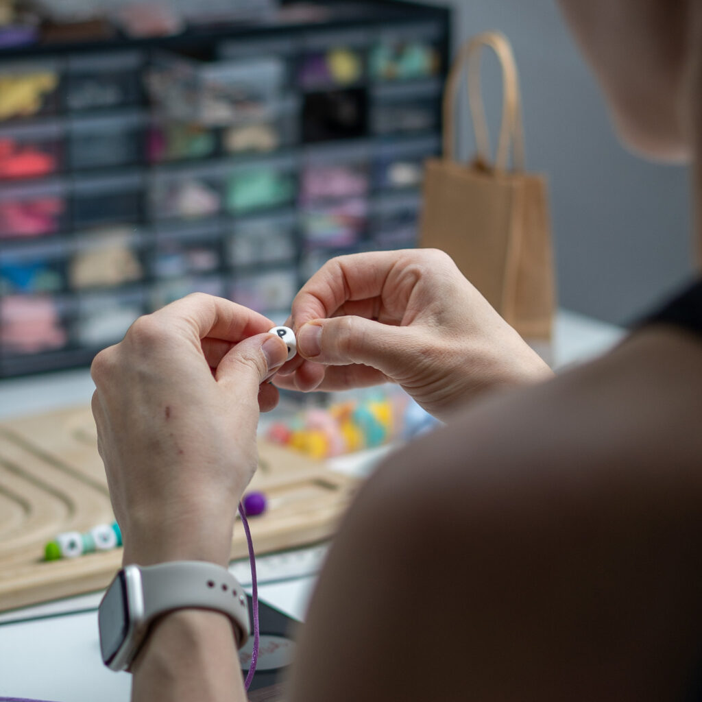 Primer plano de las manos de la artesana Raquel montando a mano un chupetero personalizado con letras de silicona blanca y cordón de seguridad en su taller.