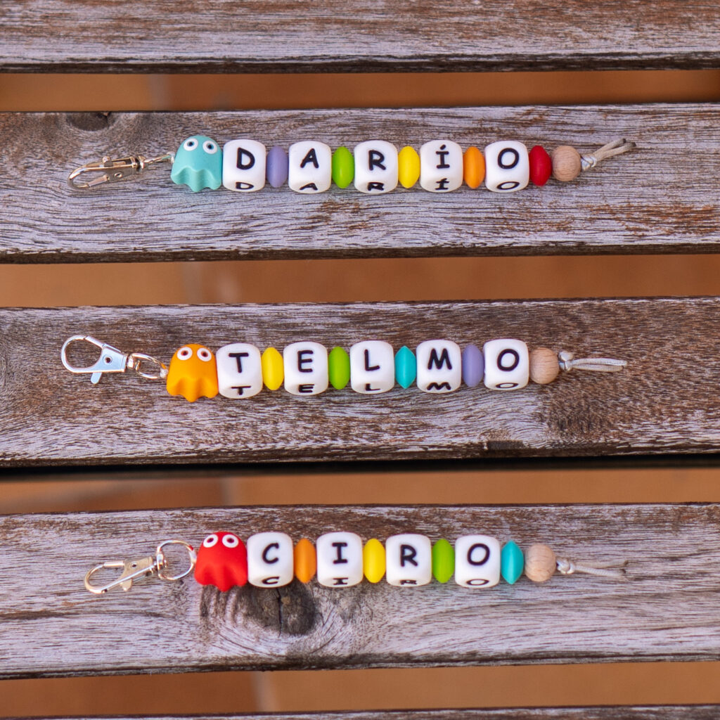 Tres llaveros con nombre personalizados (Darío, Telmo y Ciro) sobre una superficie de madera. Cuentan con un cierre de mosquetón metálico, letras cuadradas, cuentas separadoras de colores y figuras de fantasmas en silicona alimentaria.